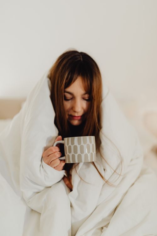 Free A woman with long hair enjoys a cozy moment wrapped in a blanket, sipping from a ceramic mug. Stock Photo