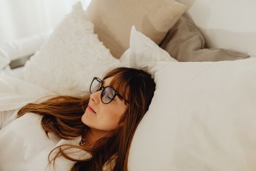 Free A calm woman wearing eyeglasses lying peacefully in a cozy bed with white pillows. Stock Photo