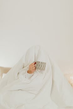 A person hides under a white blanket holding a ceramic mug, creating a cozy morning vibe.