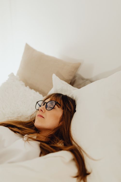 Free A woman relaxes comfortably in bed with glasses on, surrounded by soft, fluffy pillows. Stock Photo