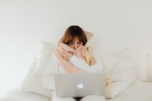 Woman wrapped in a warm blanket using a laptop on a cozy bed. Perfect for representing comfort and leisure at home.