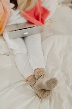 Woman relaxing on a bed with a laptop, wearing warm socks, in a cozy home setting.