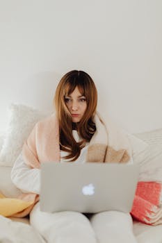 Woman in cozy setting with a laptop on her lap, wrapped in a warm blanket.