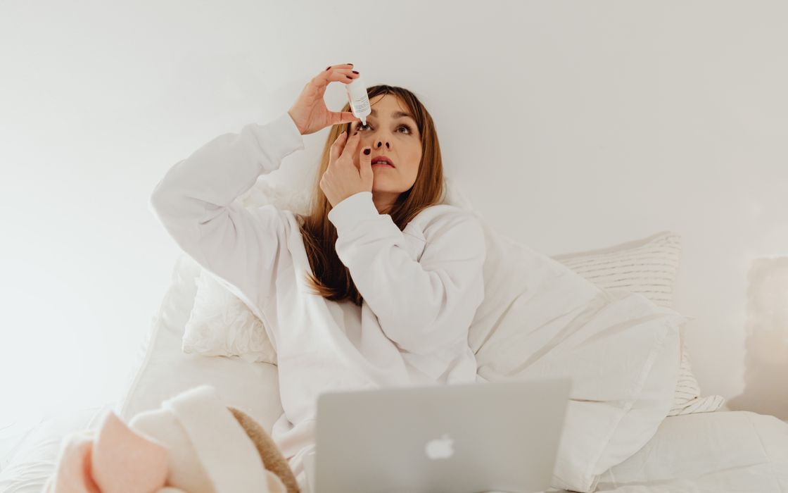 Woman applying eye drops while resting in bed with a laptop