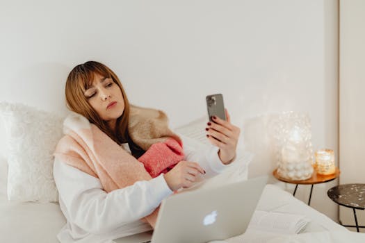 A woman wrapped in a scarf using her smartphone and laptop in a cozy bedroom setting.