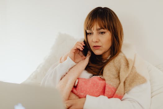 Woman in cozy setting using smartphone and laptop for remote work communication.