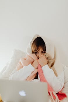 A woman wrapped in a warm blanket sits comfortably in her bedroom using a laptop.
