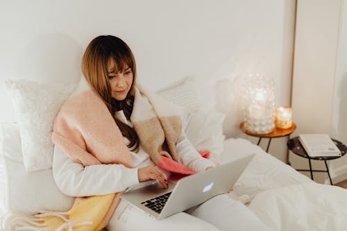 Woman in White Long Sleeves Sitting on Bed While Using a Laptop