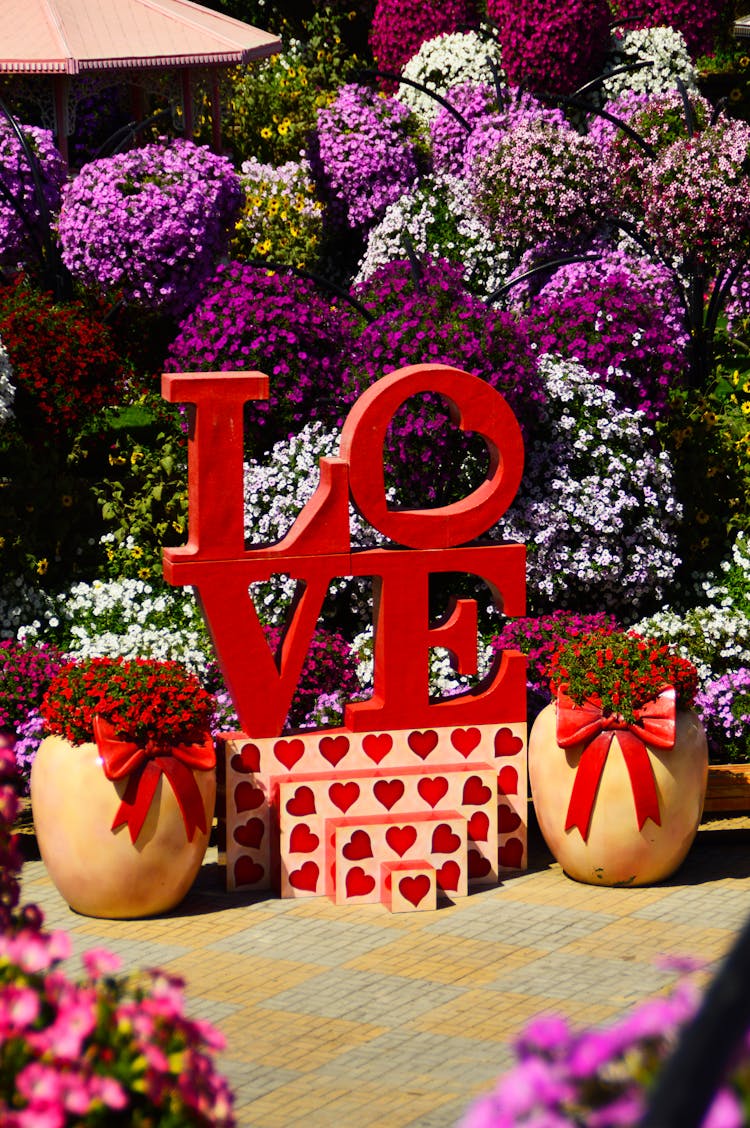 A Love Sculpture Between Potted Blooming Red Flowers In A Garden