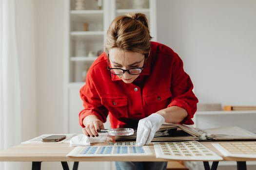 Woman in red shirt examines postage stamps with magnifying glass indoors.