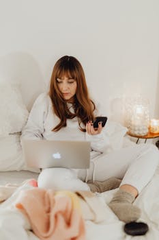 Woman multitasking with laptop and smartphone in cozy bed setting, perfect for remote work concepts.