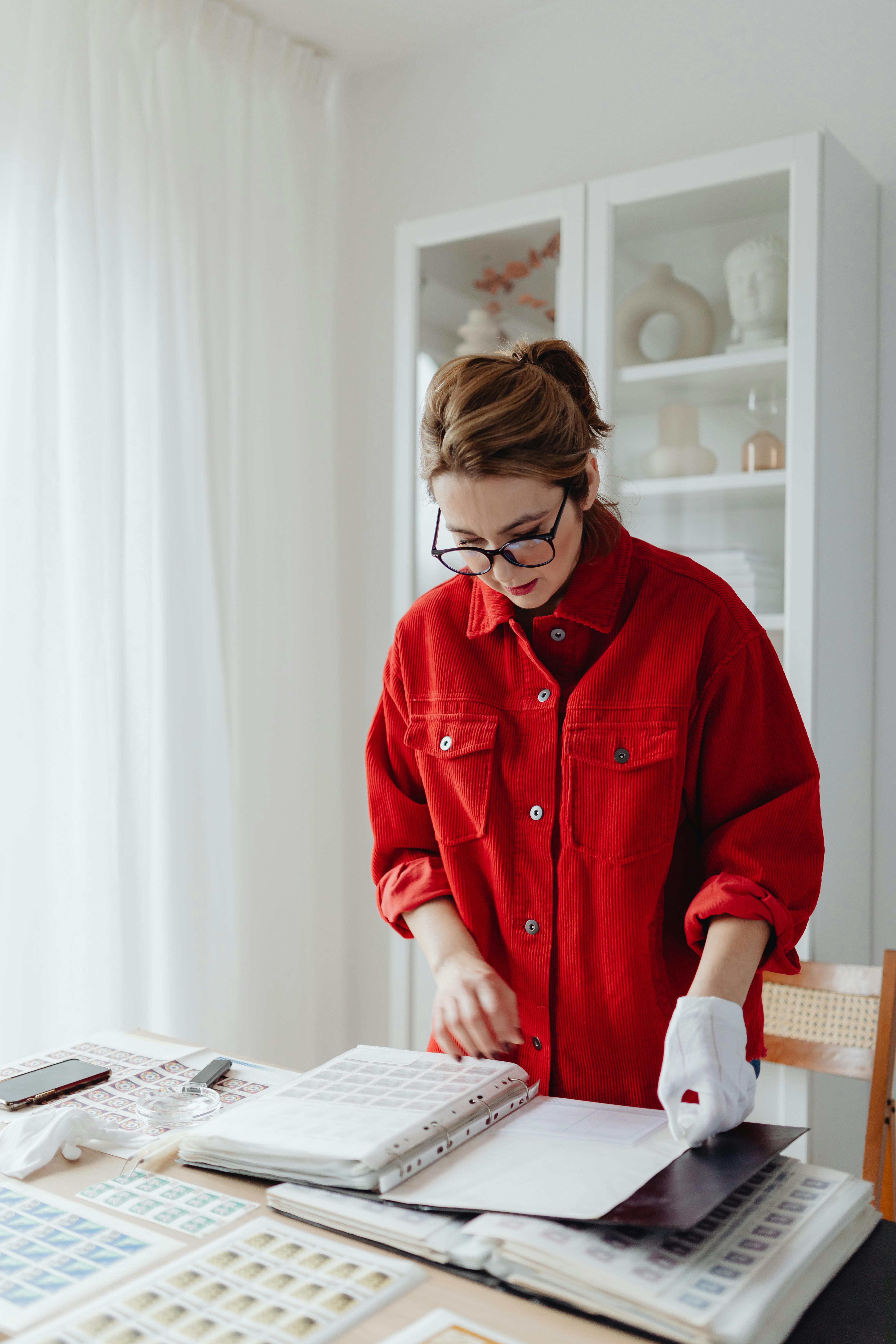 Woman in red jacket organizing a stamp collection indoors, hobby collector focused on albums.