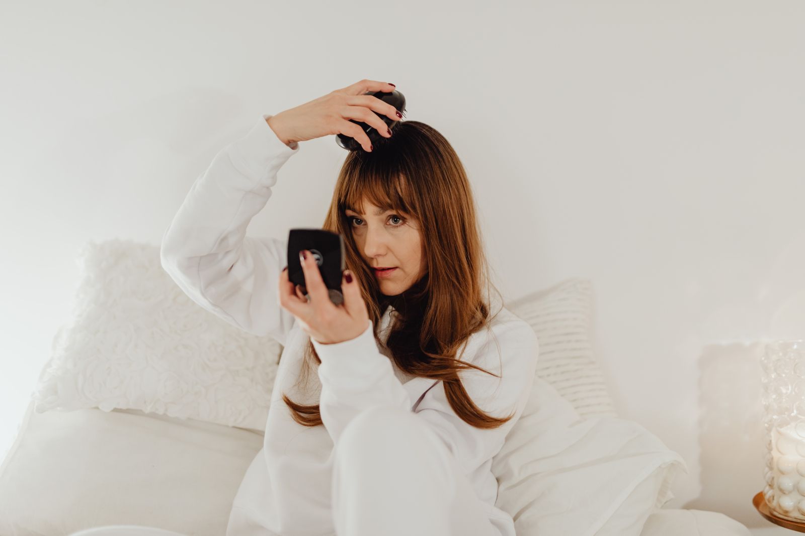 Woman with long auburn hair and bangs sits on a white bed, styling her hair with a compact mirror