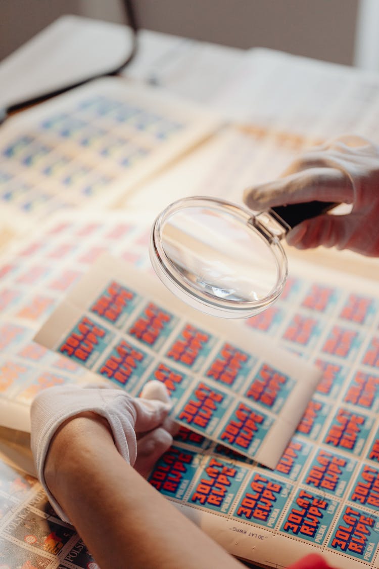 Person Holding Magnifying Glass Over Sheets Of Paper