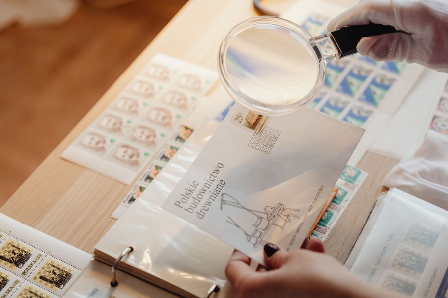Gloved hands hold a magnifying glass over postage stamps on white paper with a wooden background.