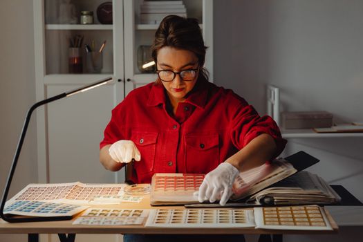 A woman carefully sorts a stamp collection under a lamp in a cozy indoor setting.