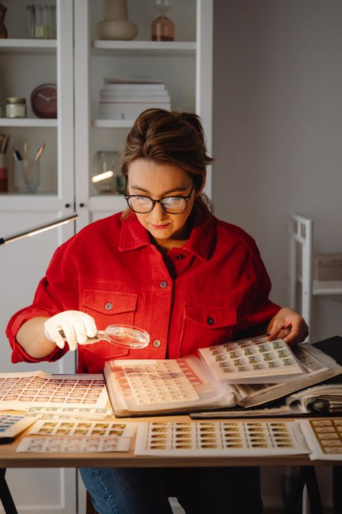 Free Woman in red jacket examining a stamp collection using a magnifying glass at home. Stock Photo
