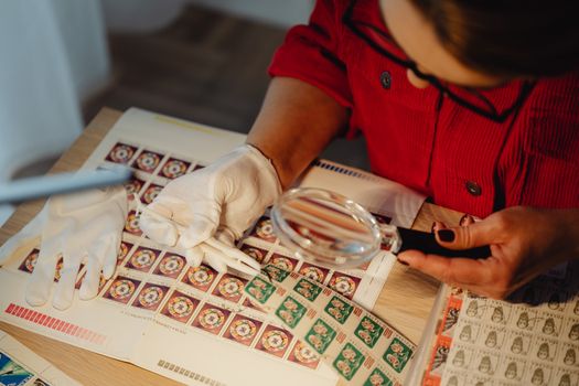 Close-up of a person with gloves inspecting colorful postage stamps using a magnifying glass on a work desk.
