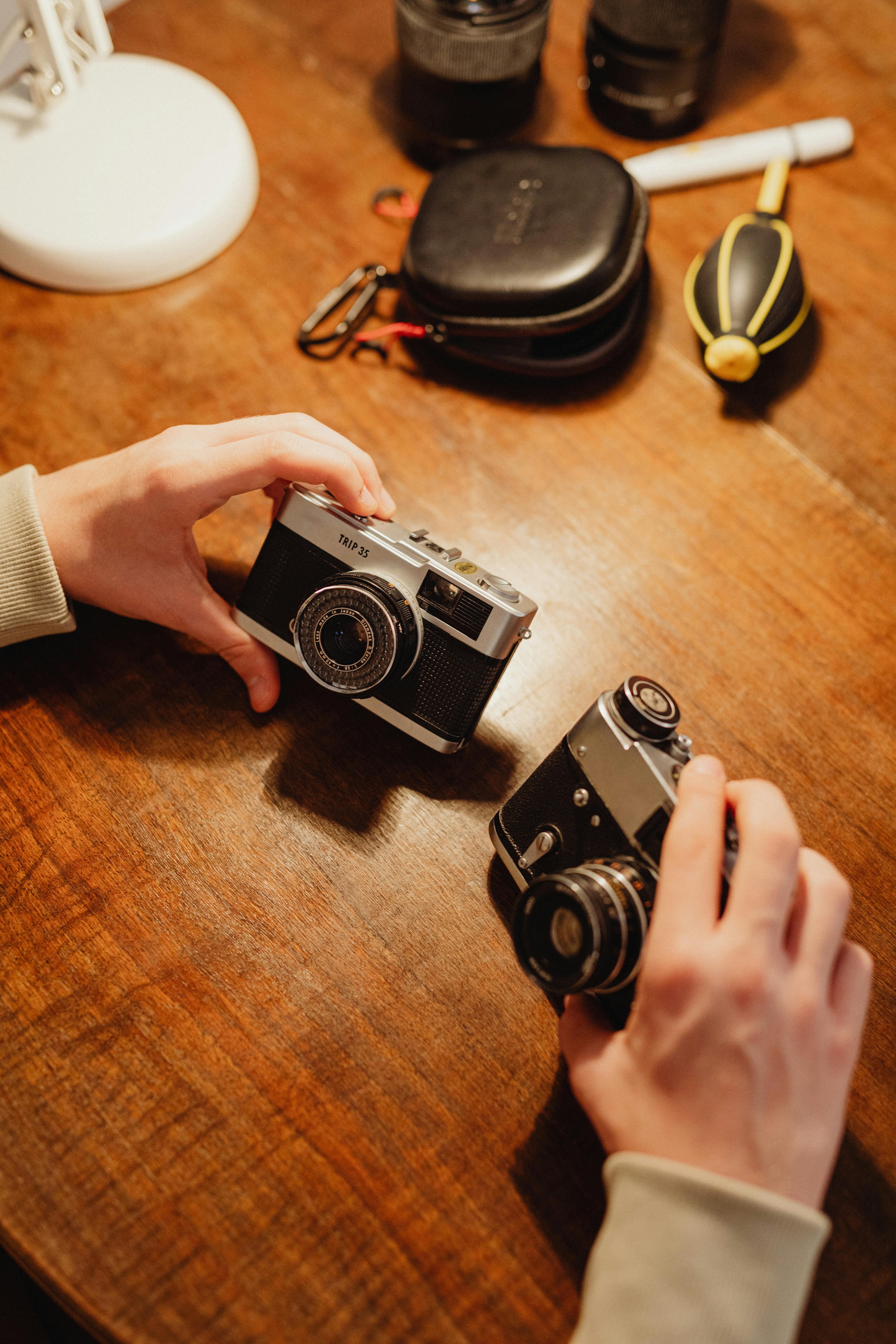 Close-up of hands holding vintage cameras on a wooden table, surrounded by photography accessories.