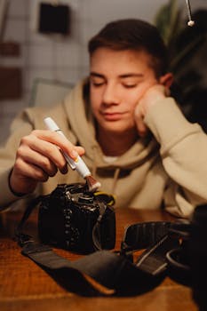 A teenager uses a cleaning brush on a camera, focusing on technology care indoors.