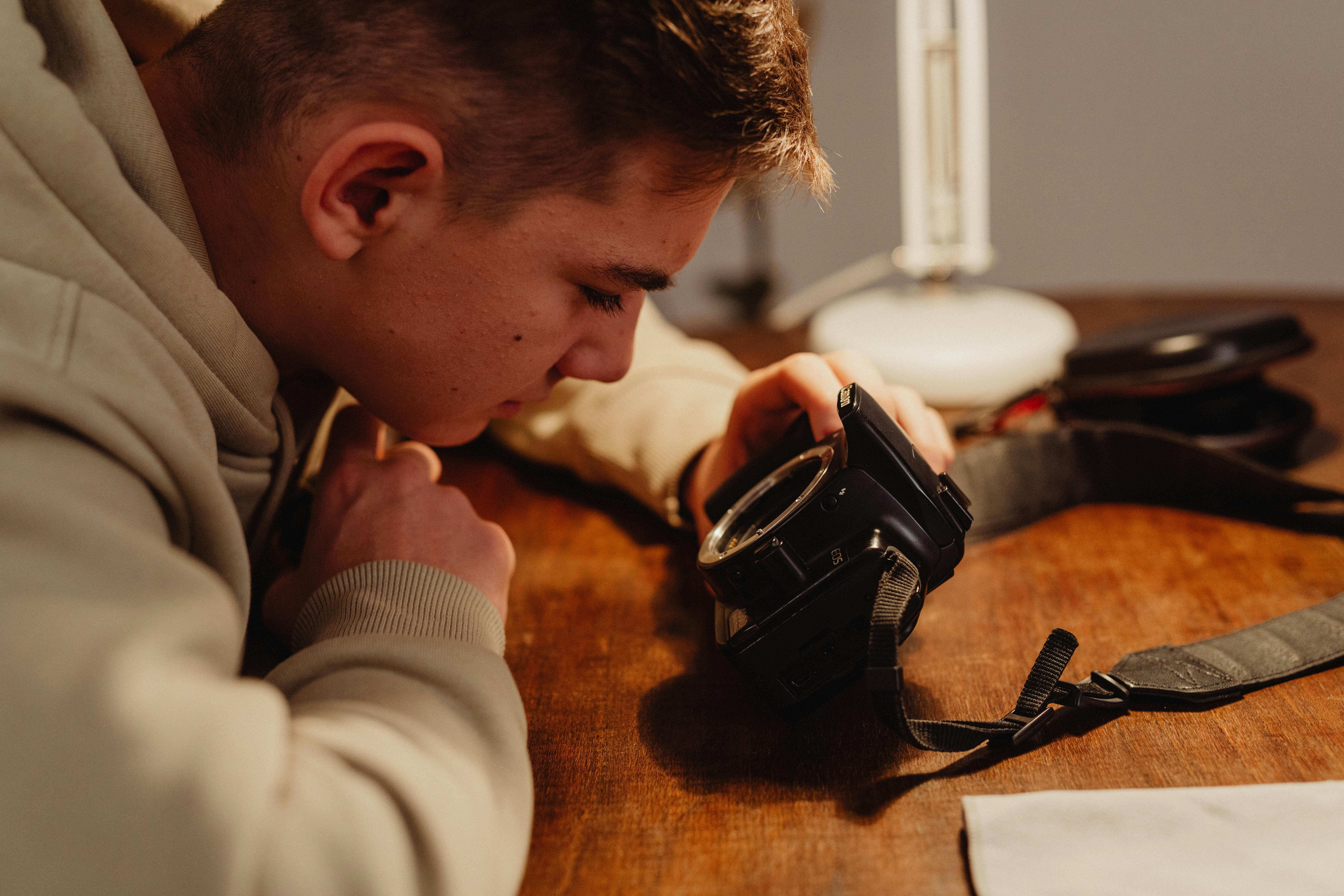 Free A young man closely inspects a vintage camera on a wooden desk indoors. Stock Photo