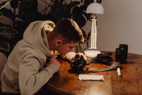 Free A young man in a hoodie examines a camera at a wooden table indoors with camera accessories. Stock Photo