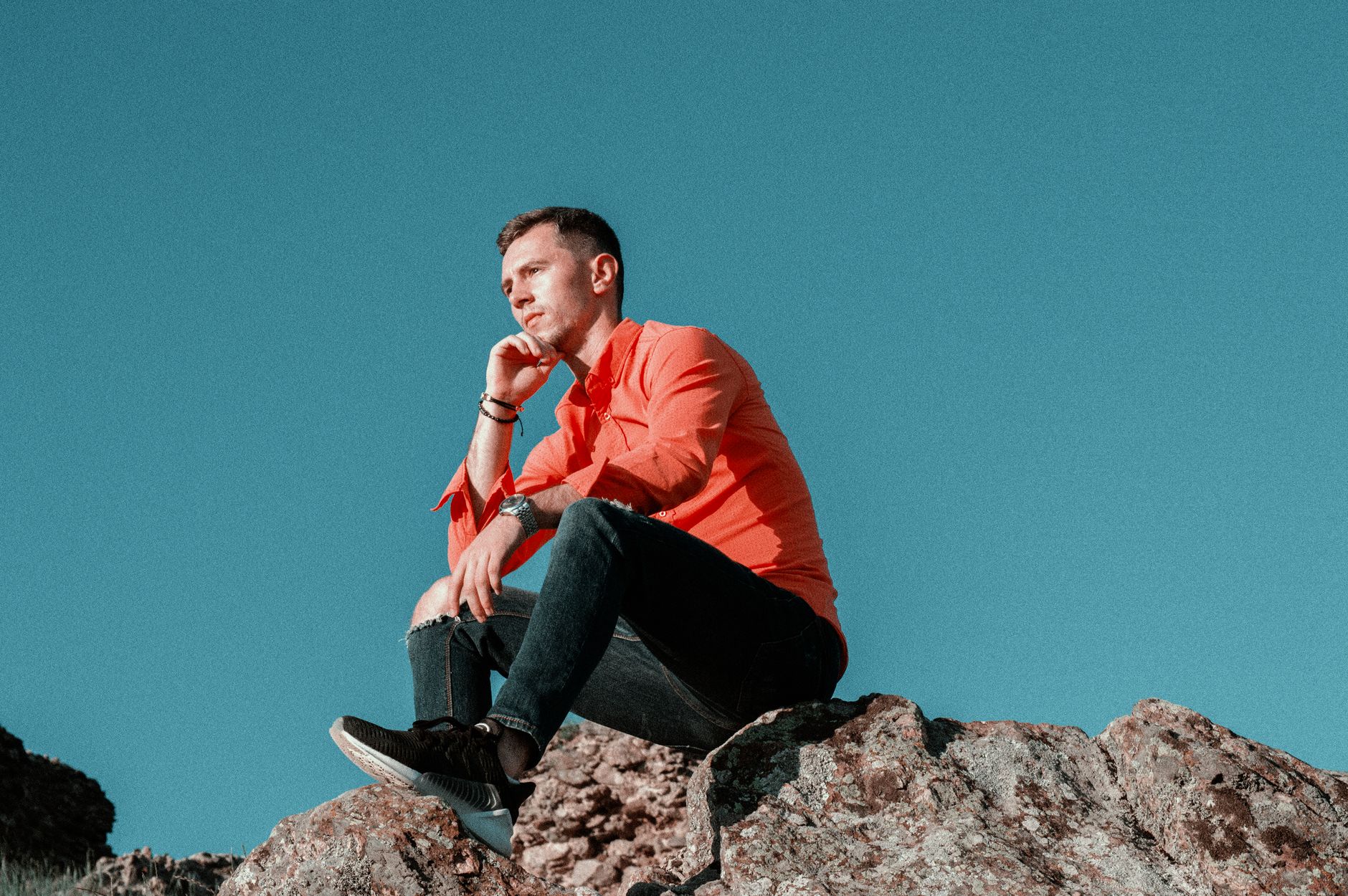 A man in a vibrant orange shirt ponders while seated on rocky terrain under a clear sky.
