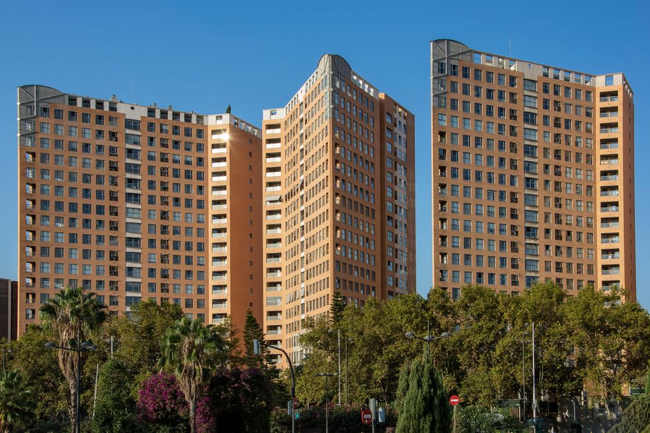 Tres modernos edificios de apartamentos altos se alzan contra un cielo azul claro, rodeados de vegetación exuberante.