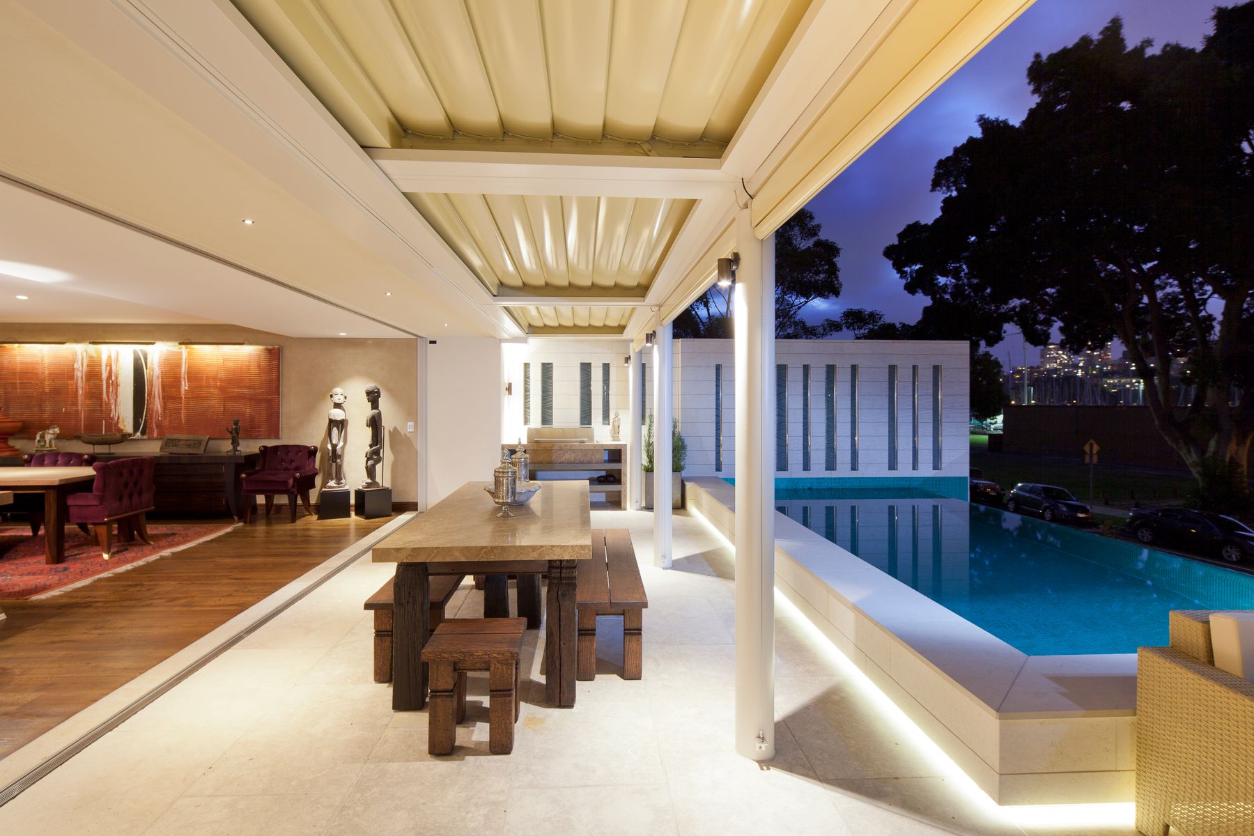 A covered terrace with a long table beside a pool at dusk