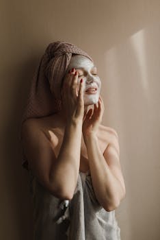 A woman enjoys a soothing facial mask at home wrapped in a towel under warm light.