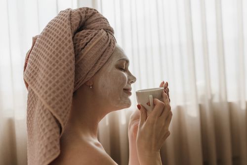 Free Woman with facial mask enjoys a cup of coffee, epitomizing relaxation and skincare. Stock Photo