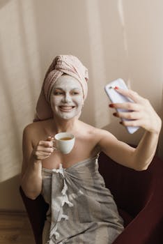 Woman with facial mask and towel taking a selfie while holding a cup of tea indoors.