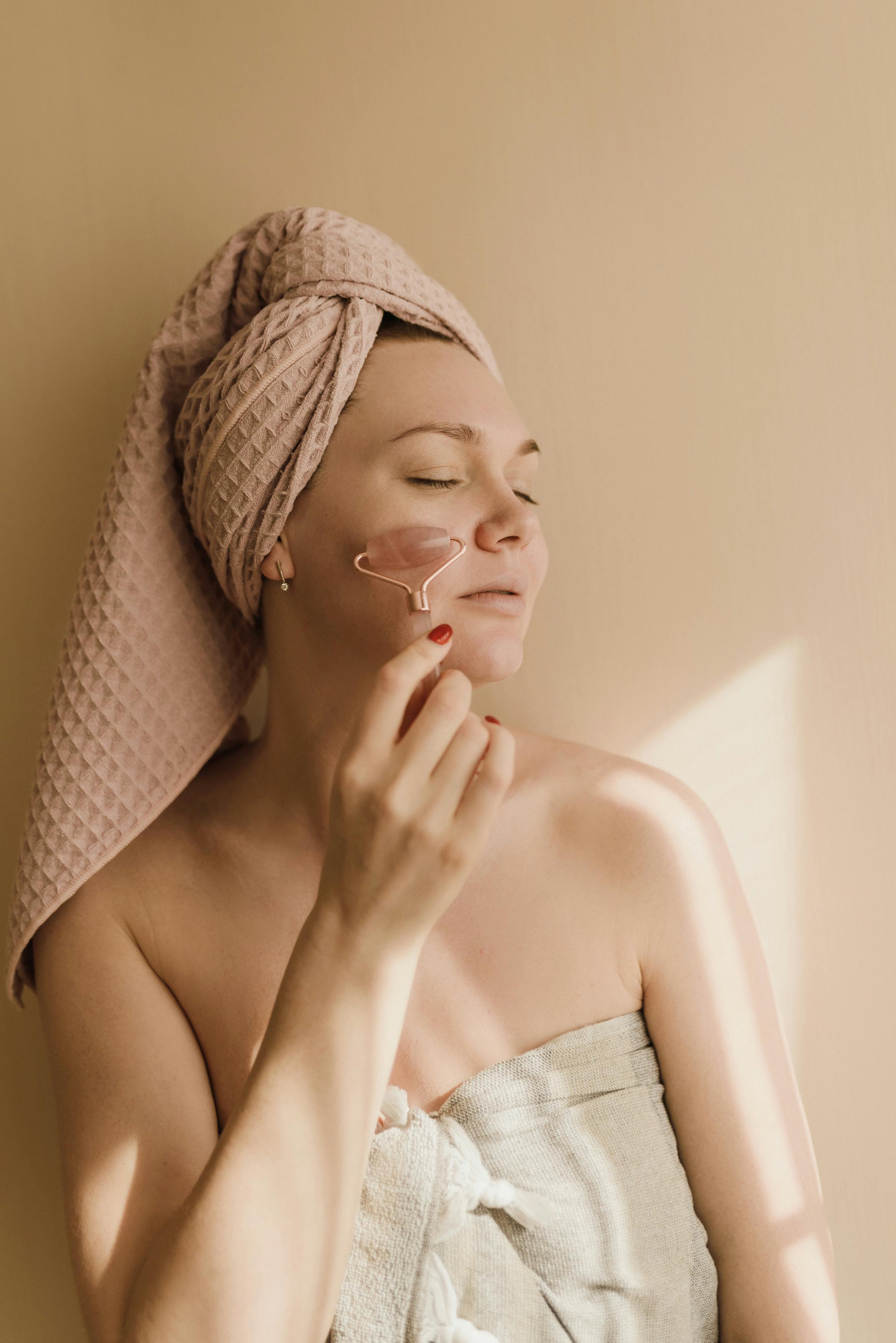 woman in white towel and pink towel on head doing facial massage with the roller