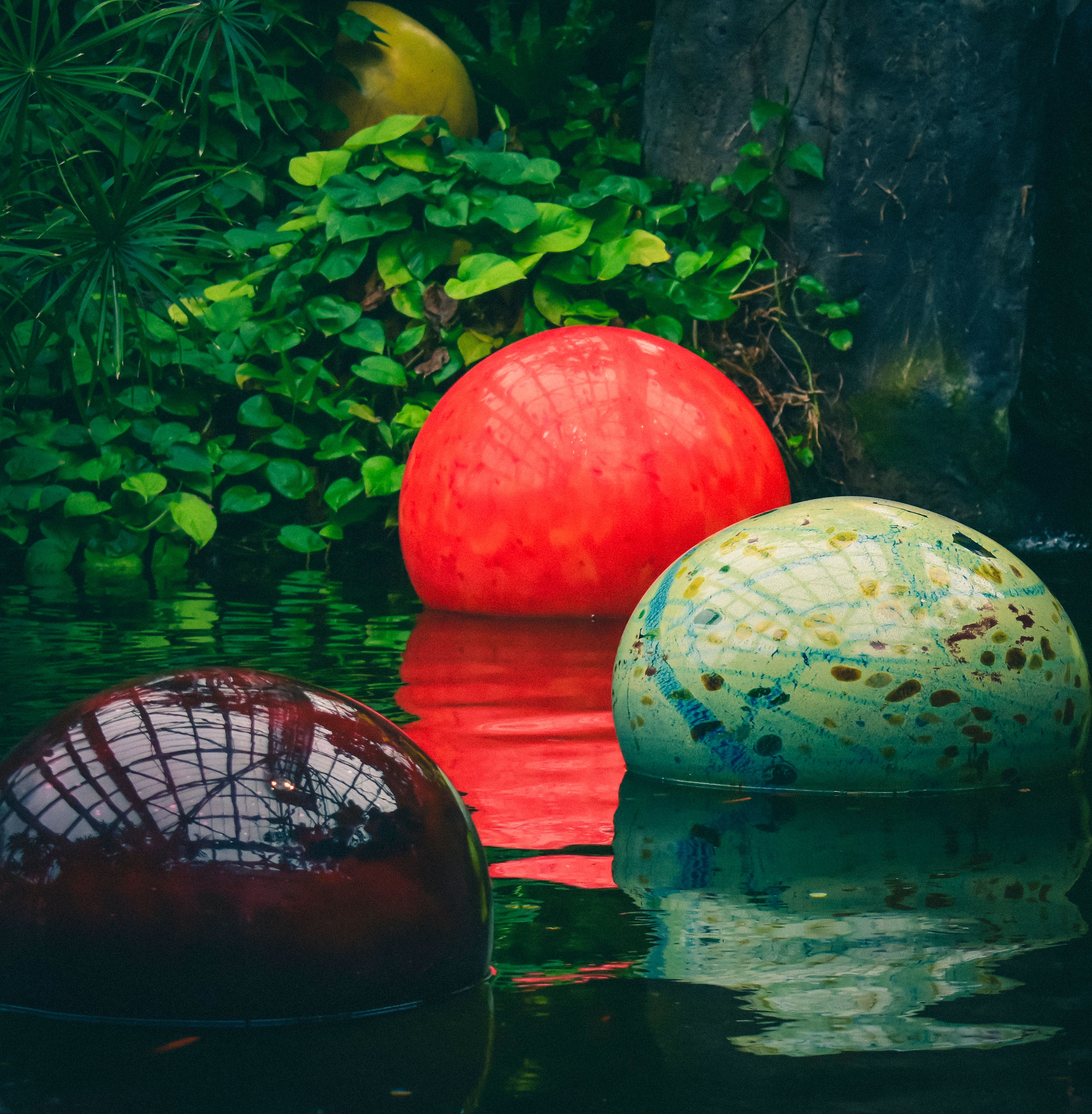 Vibrant colored spheres float in a tranquil pond surrounded by lush greenery.