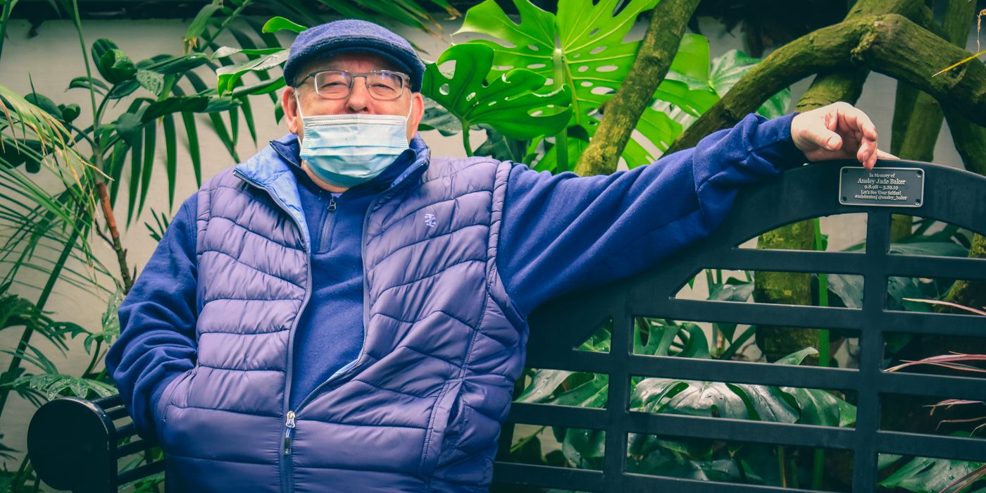Elderly man in a blue puffer jacket and cap sits outdoors, embracing new normals.