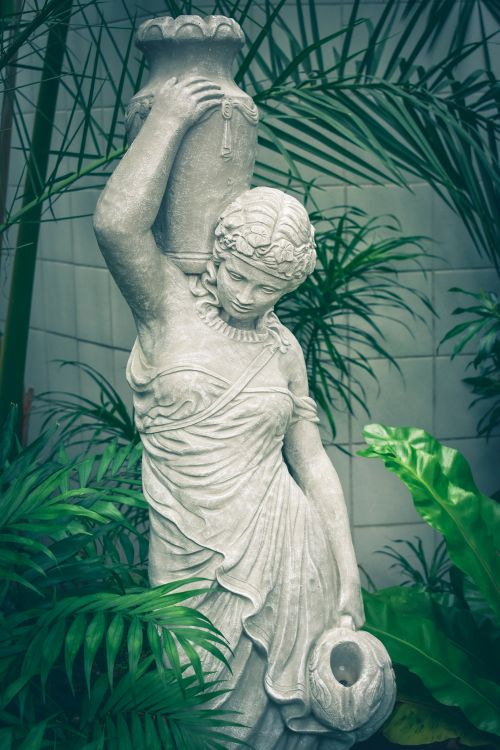 Free Elegant stone sculpture of a woman holding a jar among lush green plants. Stock Photo
