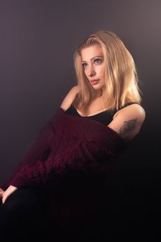 Serious female with blond hair and tattoo on shoulder wearing knitted outfit sitting on black background in modern dark studio