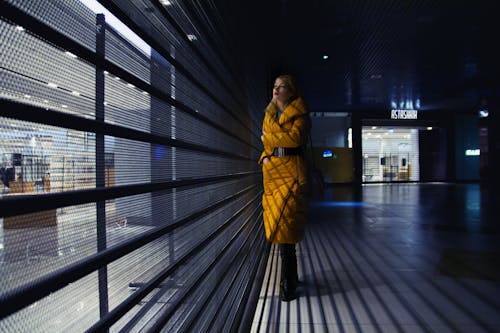 Full body of serious female in trendy outerwear standing in shadow near glass wall of contemporary building with metal partition