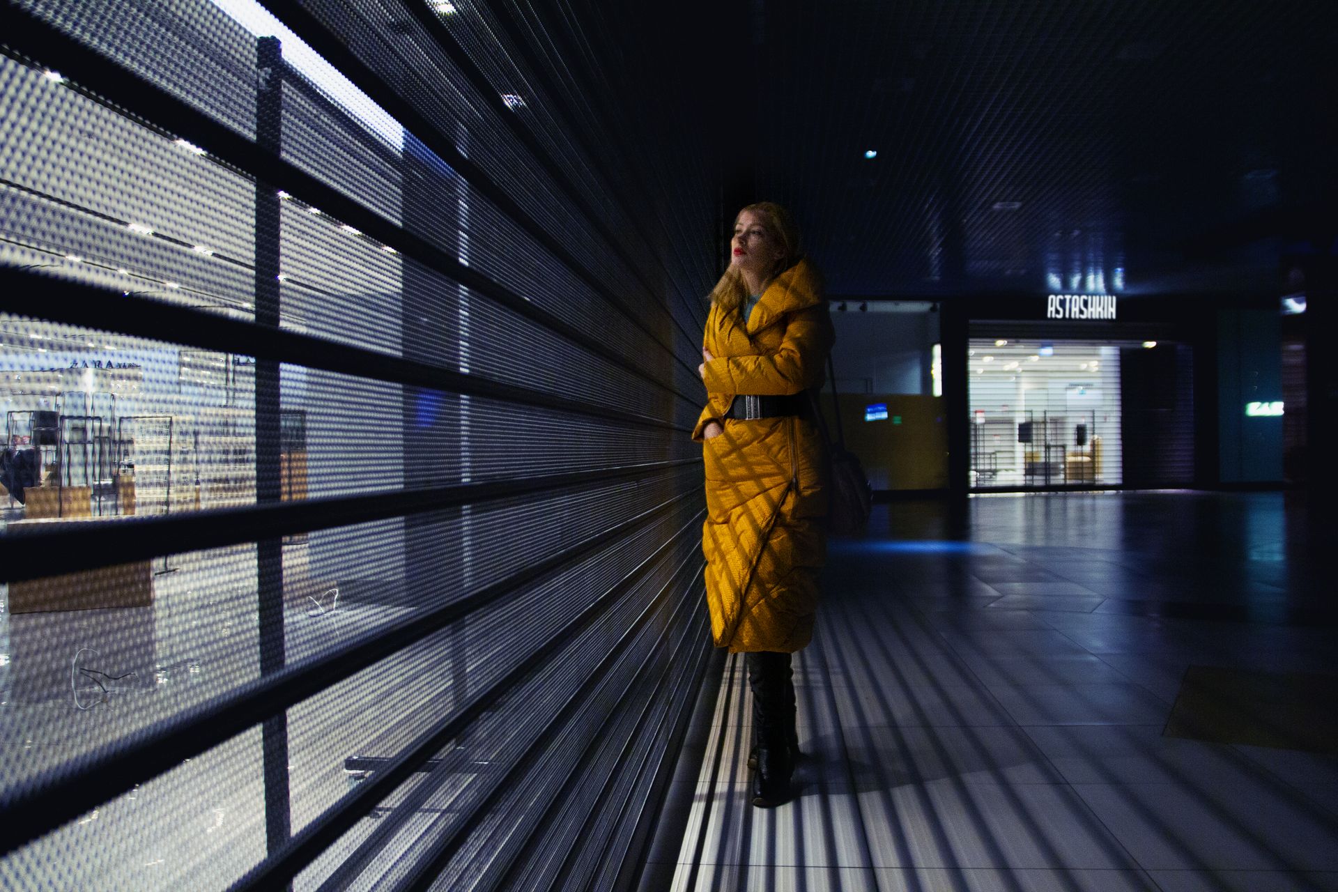 Full body of serious female in trendy outerwear standing in shadow near glass wall of contemporary building with metal partition