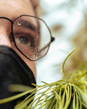 A detailed close-up of a person wearing glasses with water droplets, near green foliage.