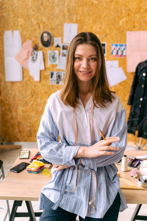 Free A smiling female tailor standing in a fashion design studio with sewing tools and sketches around. Stock Photo