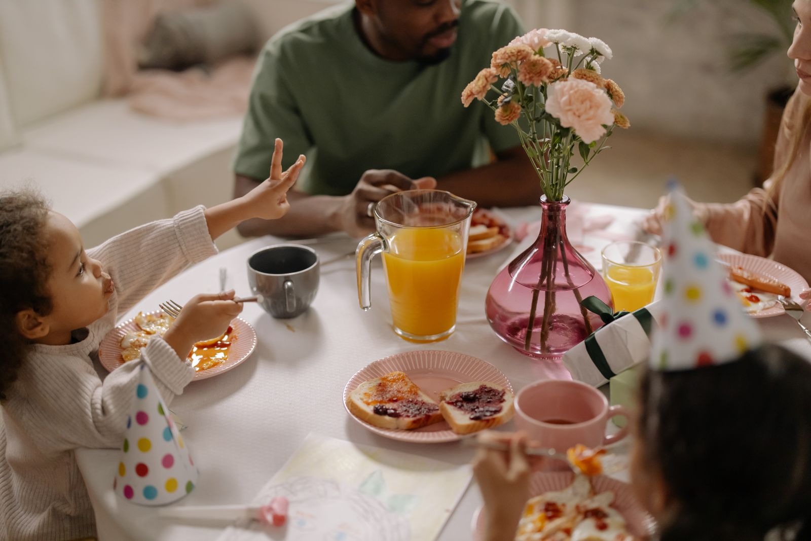 A child and several adults sit around a birthday breakfast table with plates of toast, a juice pitcher, party hats, flowers, and gifts.
