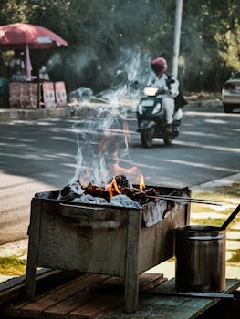 A street barbecue grill emits smoke as a motor scooter passes by on a city road.