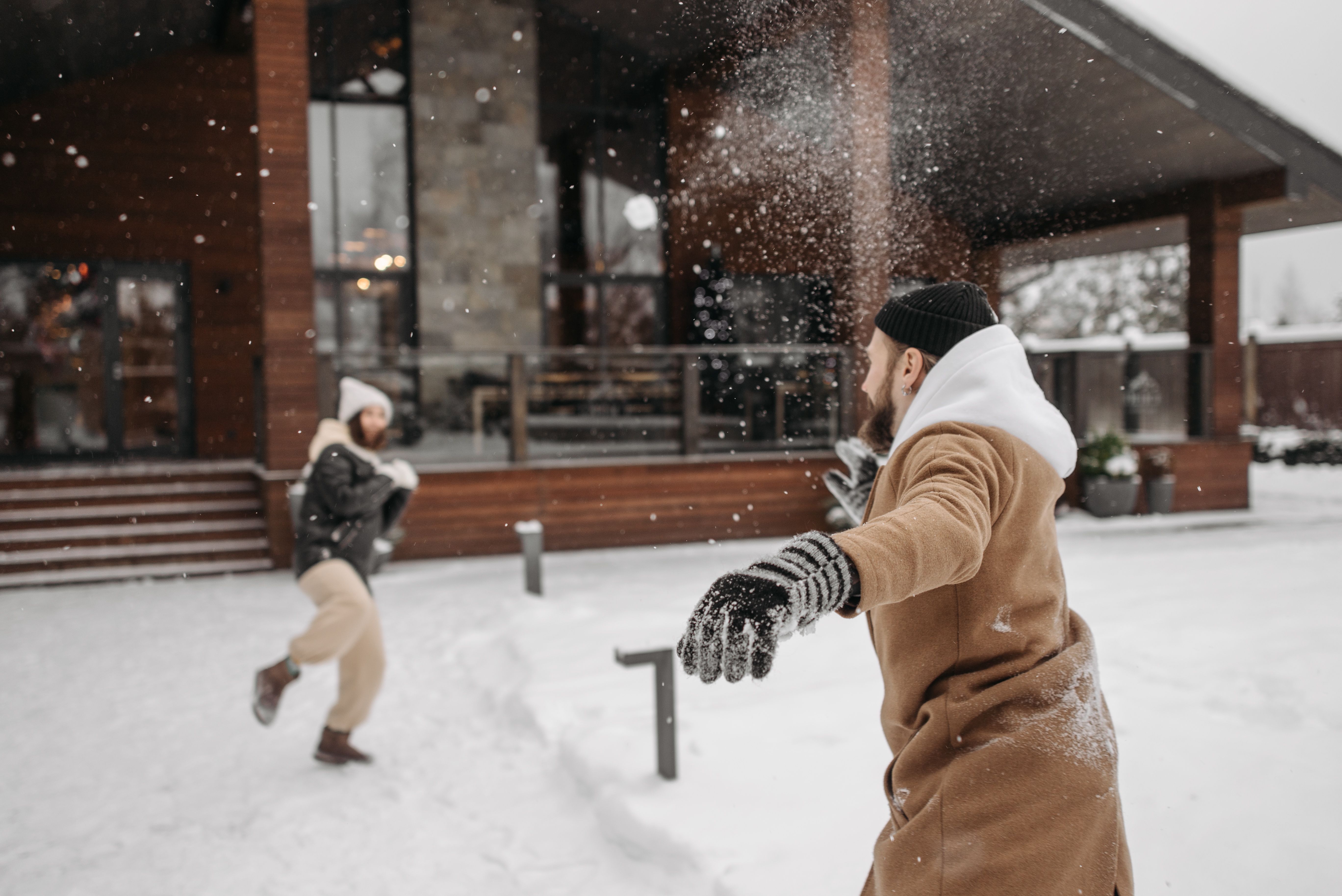 Playful couple throwing snowballs outside a cozy wooden house in wintertime.
