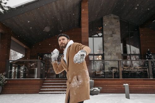 Free Bearded man with a snowball in front of a wooden cabin, enjoying winter outdoors. Stock Photo