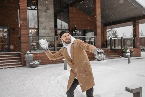 Free Bearded man in a winter coat playfully throws snowballs outside a modern cabin. Stock Photo