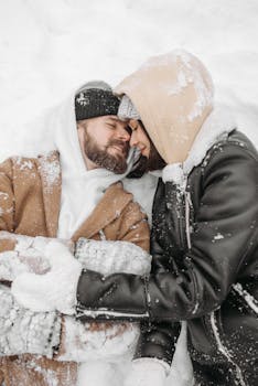 A couple embraces in winter attire, lying in snow, sharing a tender moment of love.