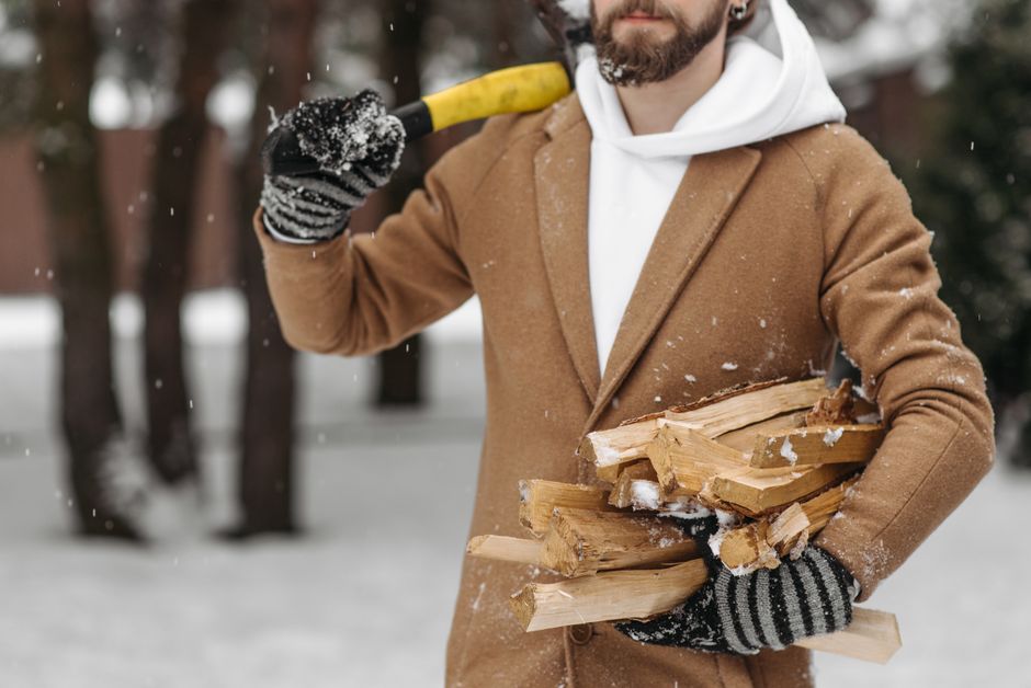 雪の降る森の中で、コートと手袋を着用した男性が薪を運んでいます。