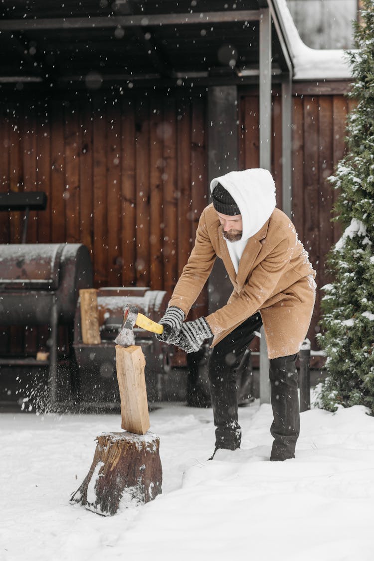 A Man In Brown Coat Standing On A Snow Covered Ground While Chopping A Wood Using An Axe