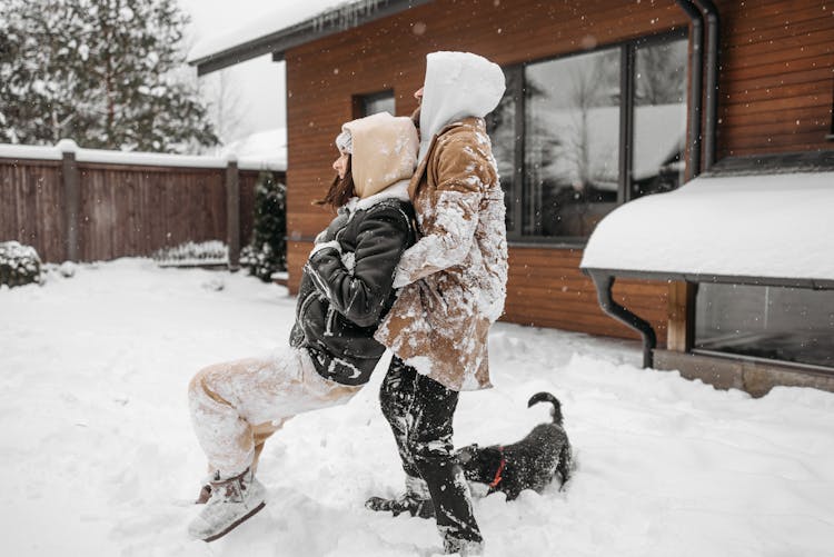 A Side View Of A Couple In Winter Clothes Playing On A Snow Covered Ground With Their Dog